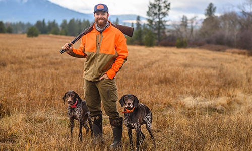 Man in orange & brown jacket in field with two dogs