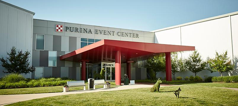 The entrance to the Purina Event Center, featuring a modern building facade with a red canopy, surrounded by well-maintained landscaping and dog sculptures on the lawn.