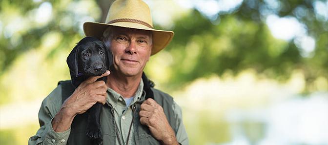 An older man wearing a straw hat and outdoor clothing holds a black puppy close to his chest. They are outdoors, with a blurred background of trees and a body of water.