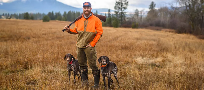 A man in an orange and brown jacket stands in a grassy field with two hunting dogs by his side, holding a rifle over his shoulder. Mountains and trees are visible in the background, creating a rugged, outdoor scene.